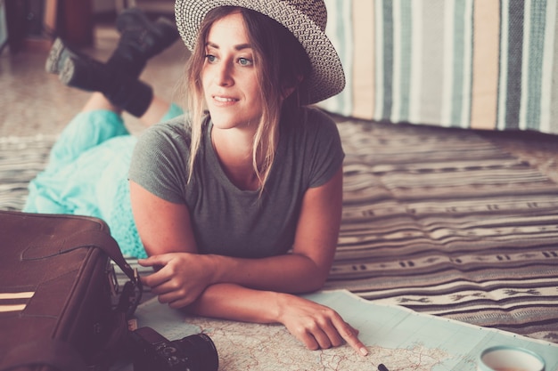 Beautiful young woman lying on carpet and pointing on atlas. Young woman in hat planning for location on map for vacation. Thoughtful woman daydreaming her travel plans while pointing on map