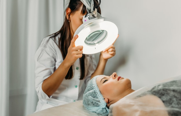 beautiful young woman lying with closed eyes and beautician looking through magnifying lamp at spa