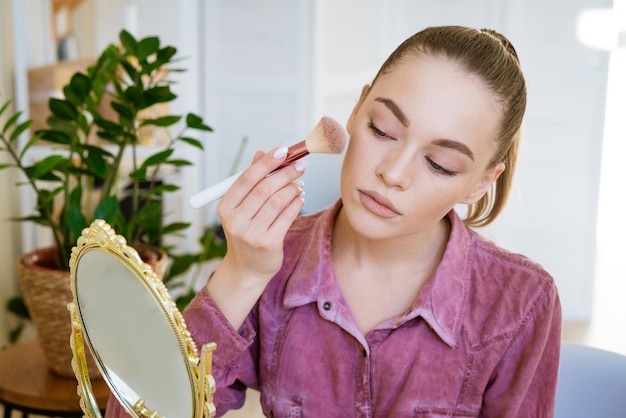 Beautiful young woman makes blush on her face using makeup brush while sitting at home in a room at ...