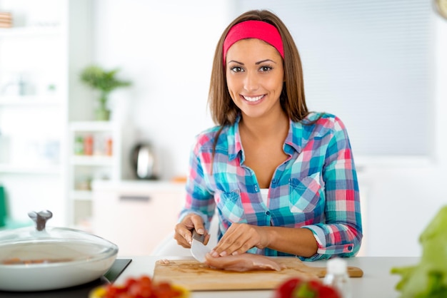 Beautiful young woman making healthy meal in the domestic kitchen. She is cutting chicken meat on the kitchen board. Looking at camera.