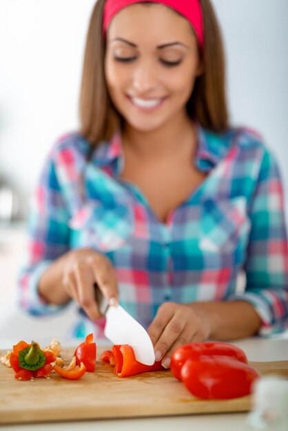 Beautiful young woman making healthy meal in the domestic kitchen. She is cutting red pepper on the kitchen board.