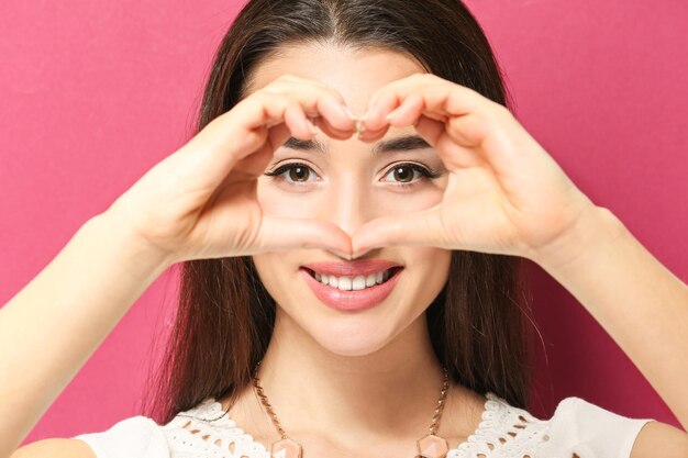 Beautiful young woman making heart with her hands on color background