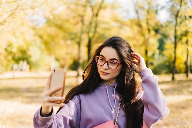 スマートフォンを使って自分撮りをし、公園で秋の天気を楽しんでいる美しい若い女性。
