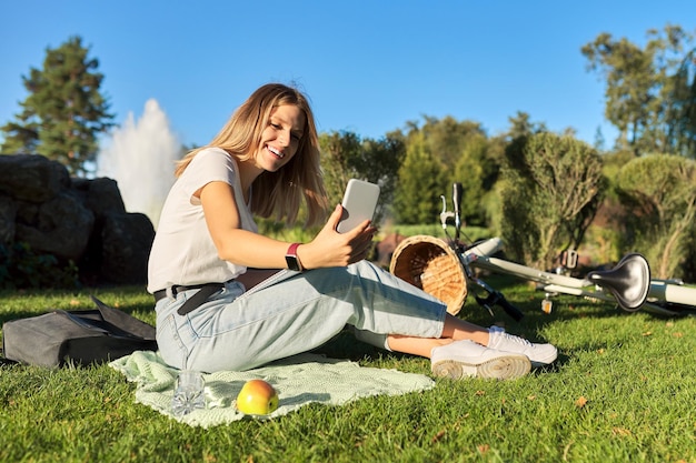 Beautiful young woman making video call on smartphone female sitting on grass in park rest online communication with friends student talking blogger blogging blog