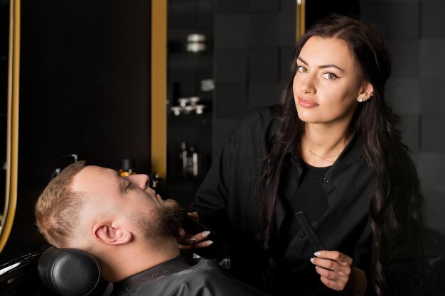 Photo beautiful young woman male master hairdresser is trimming man's beard with scissors in barber shop