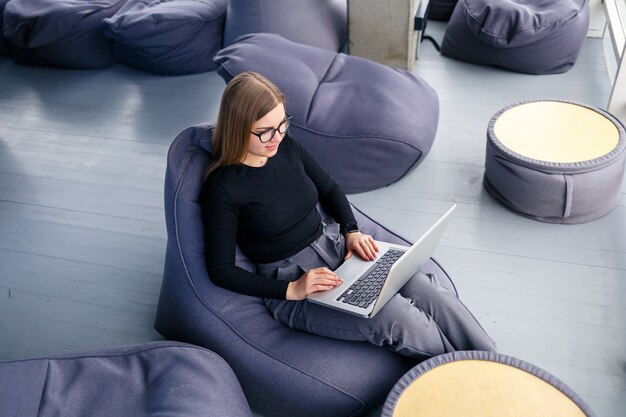 Photo a beautiful young woman manager sits with a laptop on a soft pouf near the panoramic window. girl businessman working on a new project