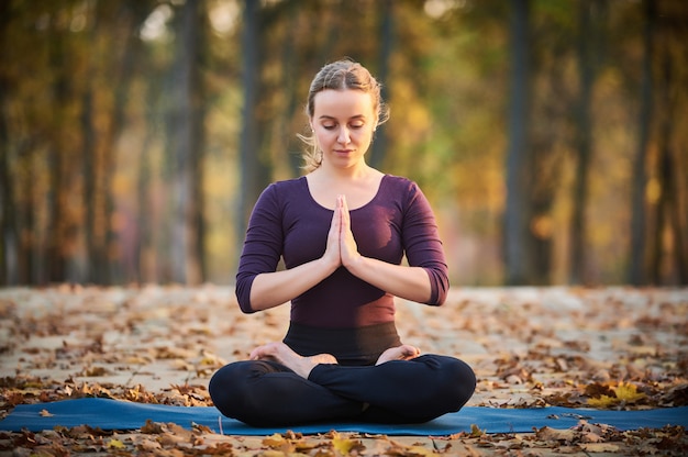 Beautiful young woman meditates in yoga asana Padmasana - Lotus pose on the wooden deck in the autumn park.