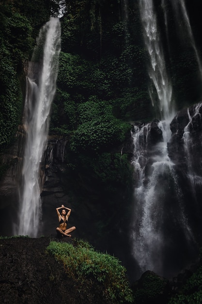 Beautiful young woman meditating in lotus position while doing yoga in a wonderful forest near waterfall. Beautiful female Practicing Yoga on Rock near Tropical Waterfall