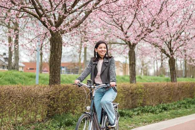 Bella giovane donna di minoranza di razze miste a cavallo in bicicletta in primavera fiorita in abiti casual con un sorriso attraente. ritratto ricreativo stile di vita.