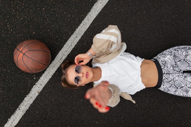 Beautiful young woman model in fashion sportswear is lying and rest on the pavement fixing her stylish sunglasses and reaching for the camera top view