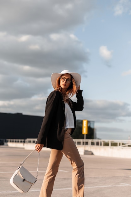 Modello di bella giovane donna in cappello di paglia in blazer nero in pantaloni alla moda in scarpe da ginnastica casual con borsa a piedi dal negozio. attraente ragazza alla moda nel parcheggio in giornata di sole.