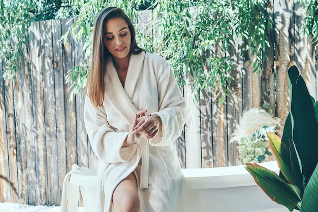 Beautiful young woman moisturizing hand cream while leaning at the bathtub outdoors