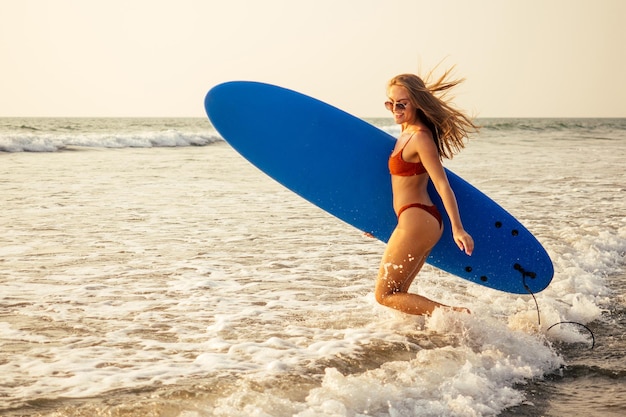 파란색 서핑 보드와 빛나는 태양 빛이 있는 주황색 비키니 수영복을 입은 아름다운 젊은 여성이 파라다이스 해변에서 물이 튀는 행복한 소녀 파도 모래와 자유와 프리랜서의 서핑 개념