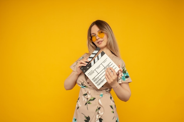 Beautiful young woman in orange eyeglasses holding classic white film making clapperboard isolated on yellow background. People sincere emotions, lifestyle concept. Advertising area