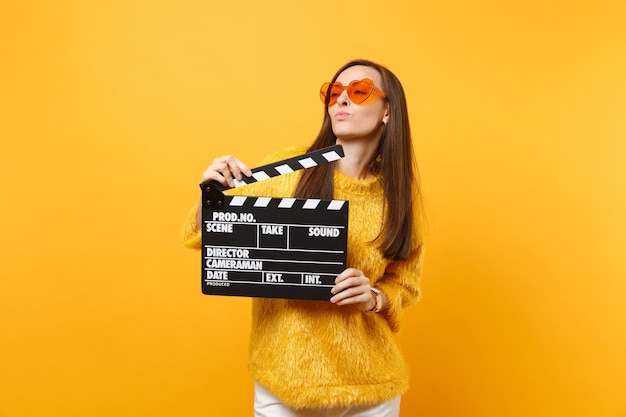 Beautiful young woman in orange heart eyeglasses looking aside and holding classic black film making clapperboard isolated on yellow background. people sincere emotions, lifestyle. advertising area
