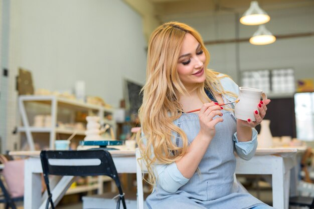 美しい若い女性が陶器のワークショップで粘土の水差しを描く