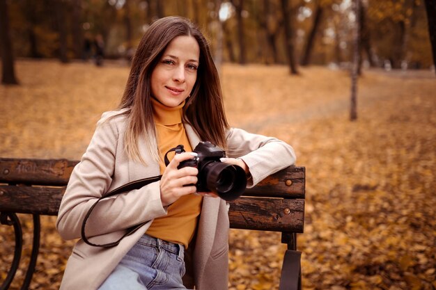 Beautiful young woman photographer with a camera in the autumn park not a bench Smiling girl enjoys the autumn weather