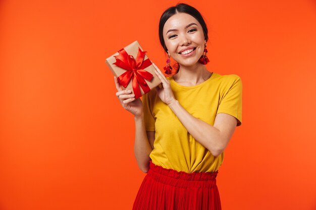 beautiful young woman posing isolated over orange wall holding present box.