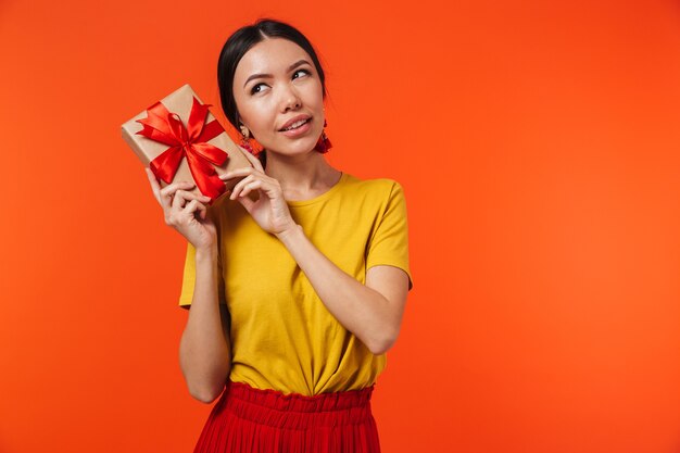 beautiful young woman posing isolated over orange wall holding present box.