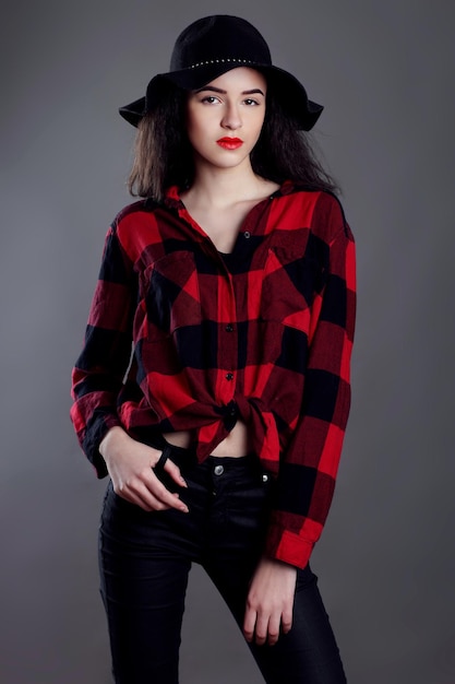 Beautiful young woman posing in red plaid shirt and black hat isolated over grey background