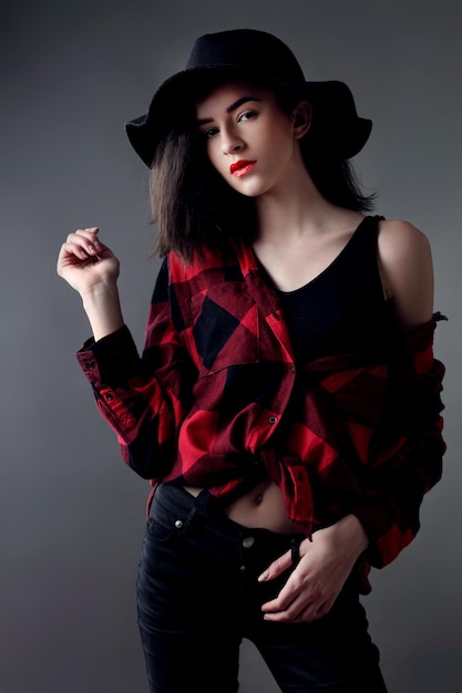 Beautiful young woman posing in red plaid shirt and black hat isolated over grey background