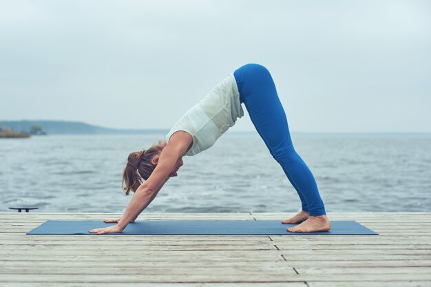 Beautiful young woman practices yoga asana downward facing dog on the wooden deck near the lake