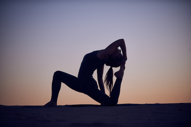 Beautiful young woman practices yoga asana King Pigeon pose rajakapotasana in the desert at night