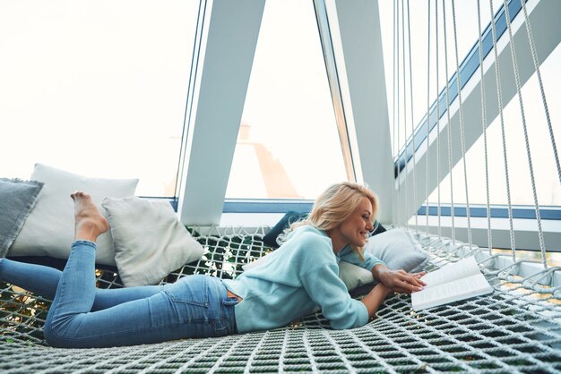 Foto bella giovane donna che legge un libro e sorride mentre si rilassa in una grande amaca a casa