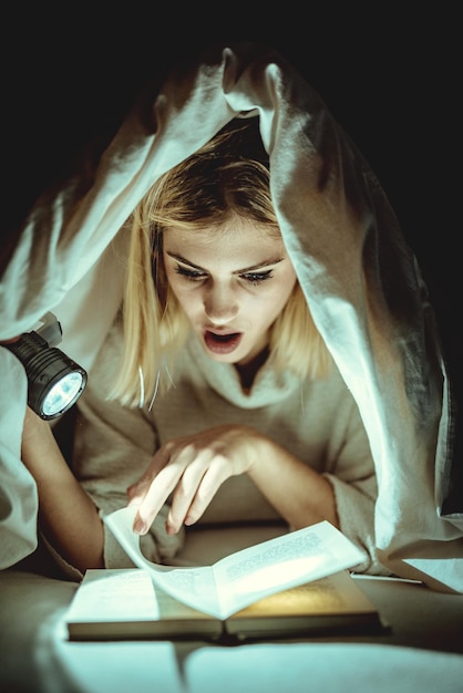 Фото Красивая молодая женщина читает книгу под одеялом и держит фонарик.
