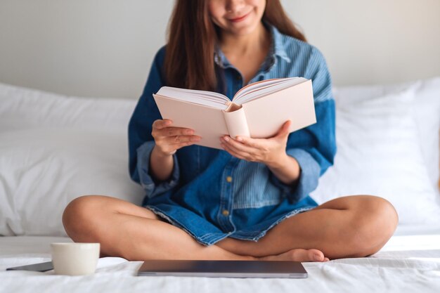 A beautiful young woman reading book while sitting on a white cozy bed at home