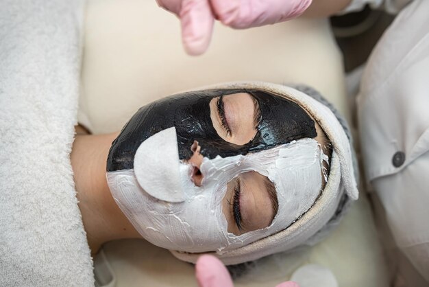 beautiful young woman receive white and black facial mask on half face lying on spa