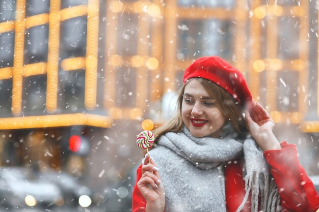 Beautiful young woman in red coat holding sweet candy during the snowfall. Empty space