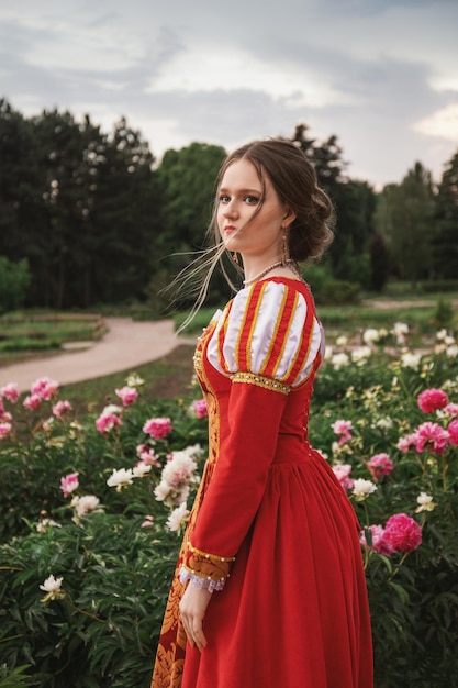 beautiful young woman in a red medieval dress is standing in the garden in the pink peonies