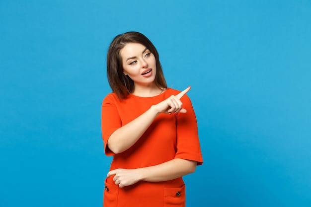 Beautiful young woman in red orange dress standing posing pointing hands fingers on workspace isolated over trendy blue wall background, studio portrait. People lifestyle concept. Mock up copy space.