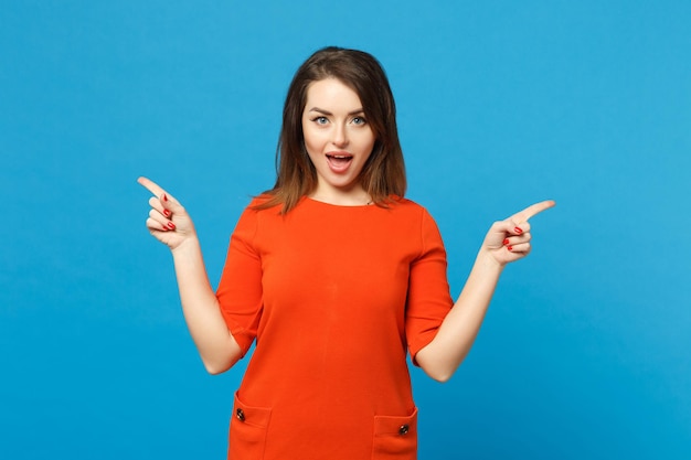 Beautiful young woman in red orange dress standing posing pointing hands fingers on workspace isolated over trendy blue wall background, studio portrait. People lifestyle concept. Mock up copy space.