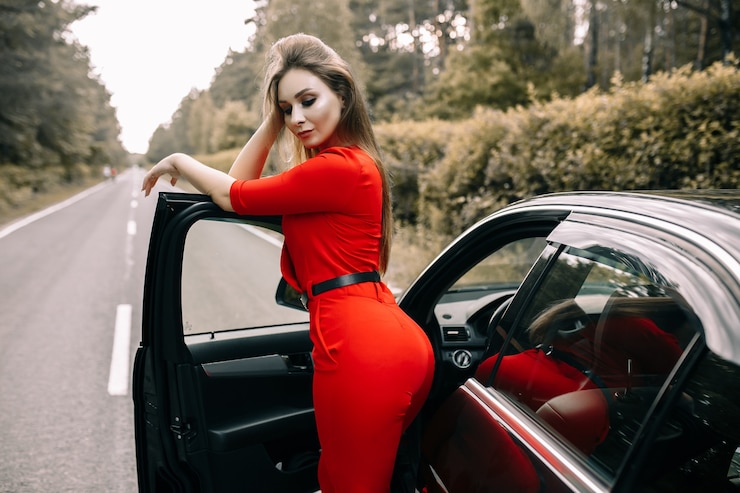 Photo 赤いオーバーオールを着た美しい若い女性が森の中の何もない道で黒い車のそばに立っています