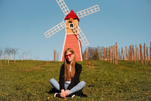 Beautiful young woman in red sunglasses sits on the grass against the backdrop of the red mill.