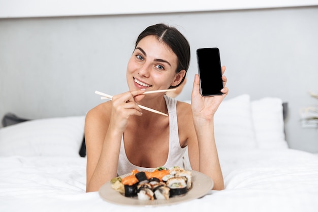 Beautiful young woman relaxing on bed at home, eating sushi from a plate, holding mobile phone