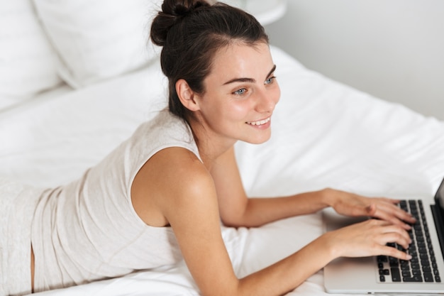 Beautiful young woman relaxing on bed at home, using laptop computer