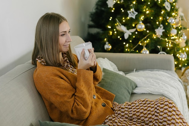 Beautiful young woman relaxing at home on Christmas day, sitting on couch near the Christmas tree, drinking tea or coffee