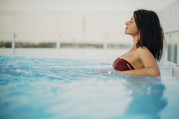 Bella giovane donna che si rilassa nella vasca idromassaggio della piscina sorridendo e godendosi la giornata felice ragazza bruna in vacanza estiva in resort di lusso che nuota in piscina sul tetto con vista sul mare copia spazio