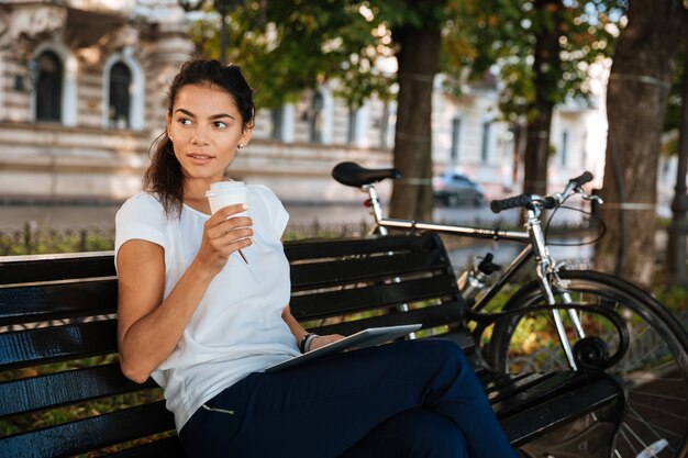 Beautiful young woman resting on the bench with cup of coffee at the city park