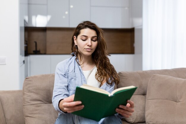 Beautiful young woman resting at home sitting on the couch and reading an interesting book