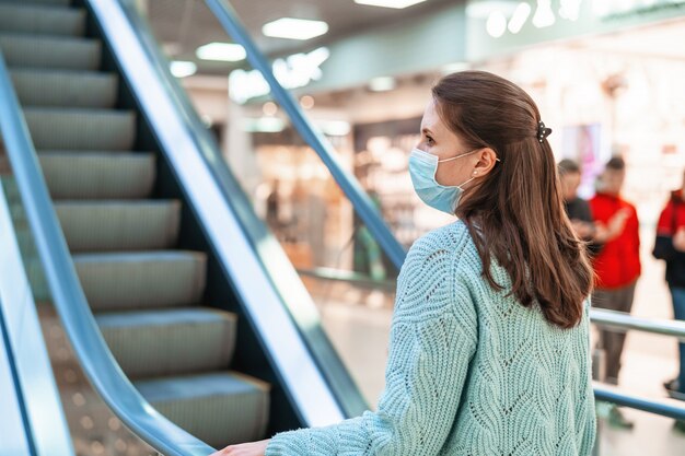 Beautiful young woman rides escalator of a shopping center with protective mask