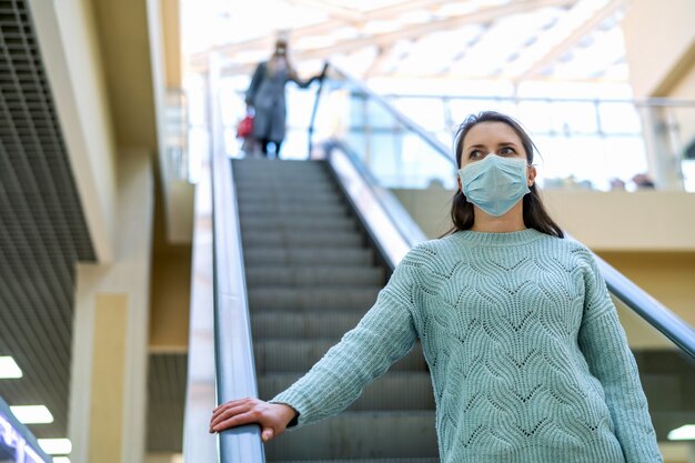 Beautiful young woman rides escalator of a shopping center with protective mask