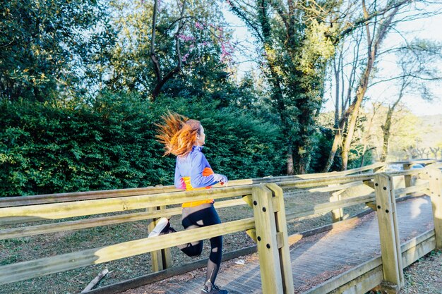 Beautiful young woman running by a park in spring or summer in\
a forest is down a walkway from behind. sport self-care\
concept