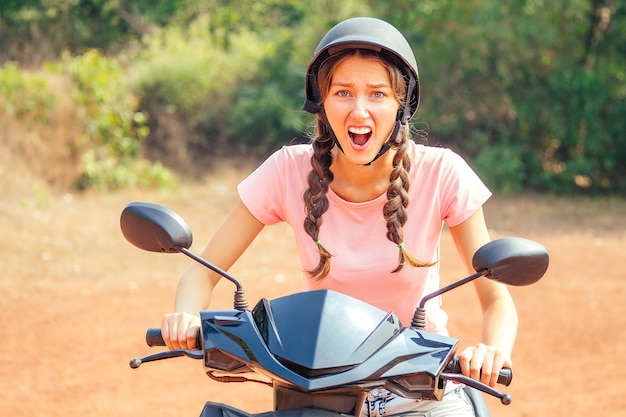 안전모를 쓴 아름답고 젊은 여성이 오토바이(자전거)에 앉아 두려움에 울부짖습니다. 스쿠터 및 사고 안전 운전의 개념