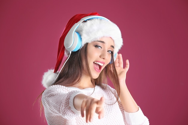 Beautiful young woman in Santa hat listening to Christmas music on color background