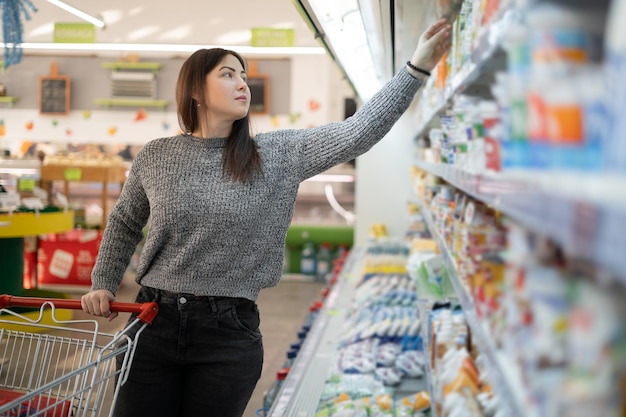 食料品店のスーパーマーケットの乳製品食料品売り場でヨーグルトを買う美しい若い女性