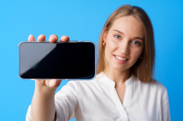 Beautiful young woman showing smartphone screen against blue background